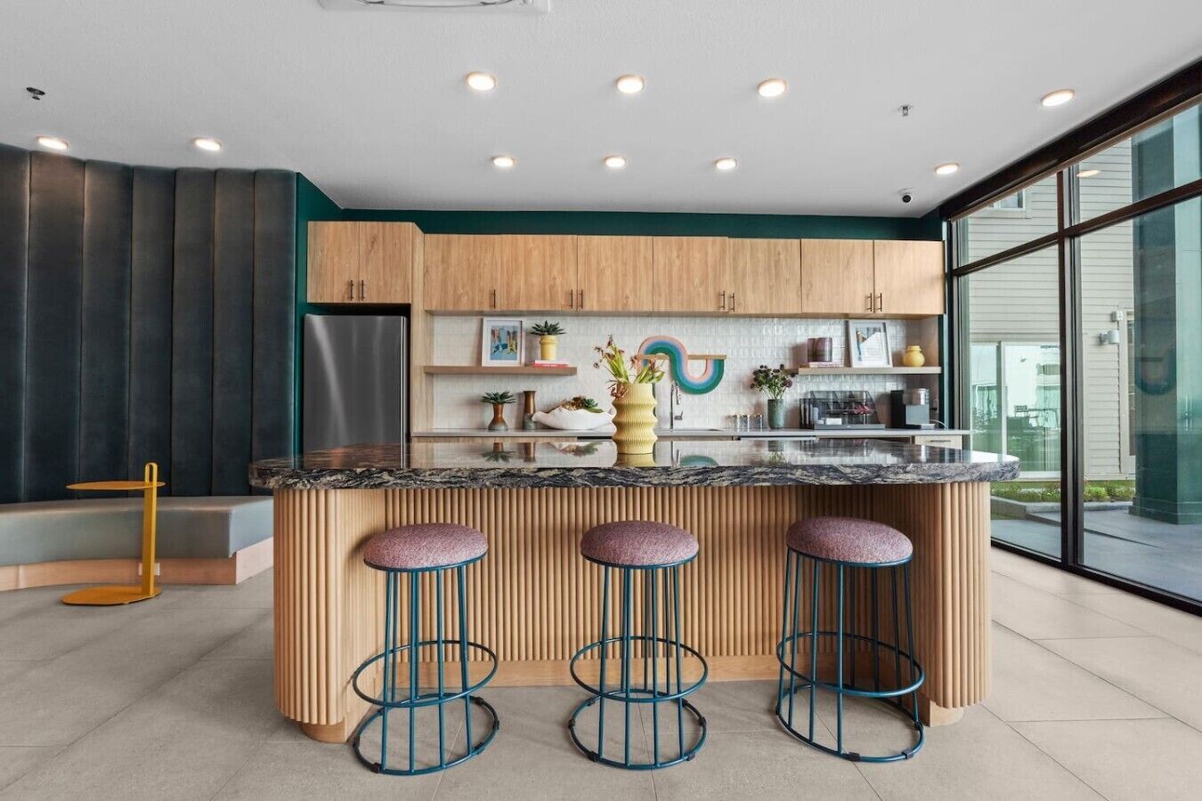A modern kitchen with a central island, bar stools, wooden cabinets, and a large window providing a view outside.