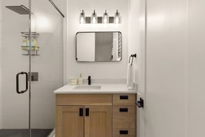 A modern bathroom with a glass shower enclosure, a white sink, wooden cabinets, a mirror with a black frame, and a towel rack. - Mountain Views | Apt Walk to Downtown | Sleeps 6 (Buena Vista)