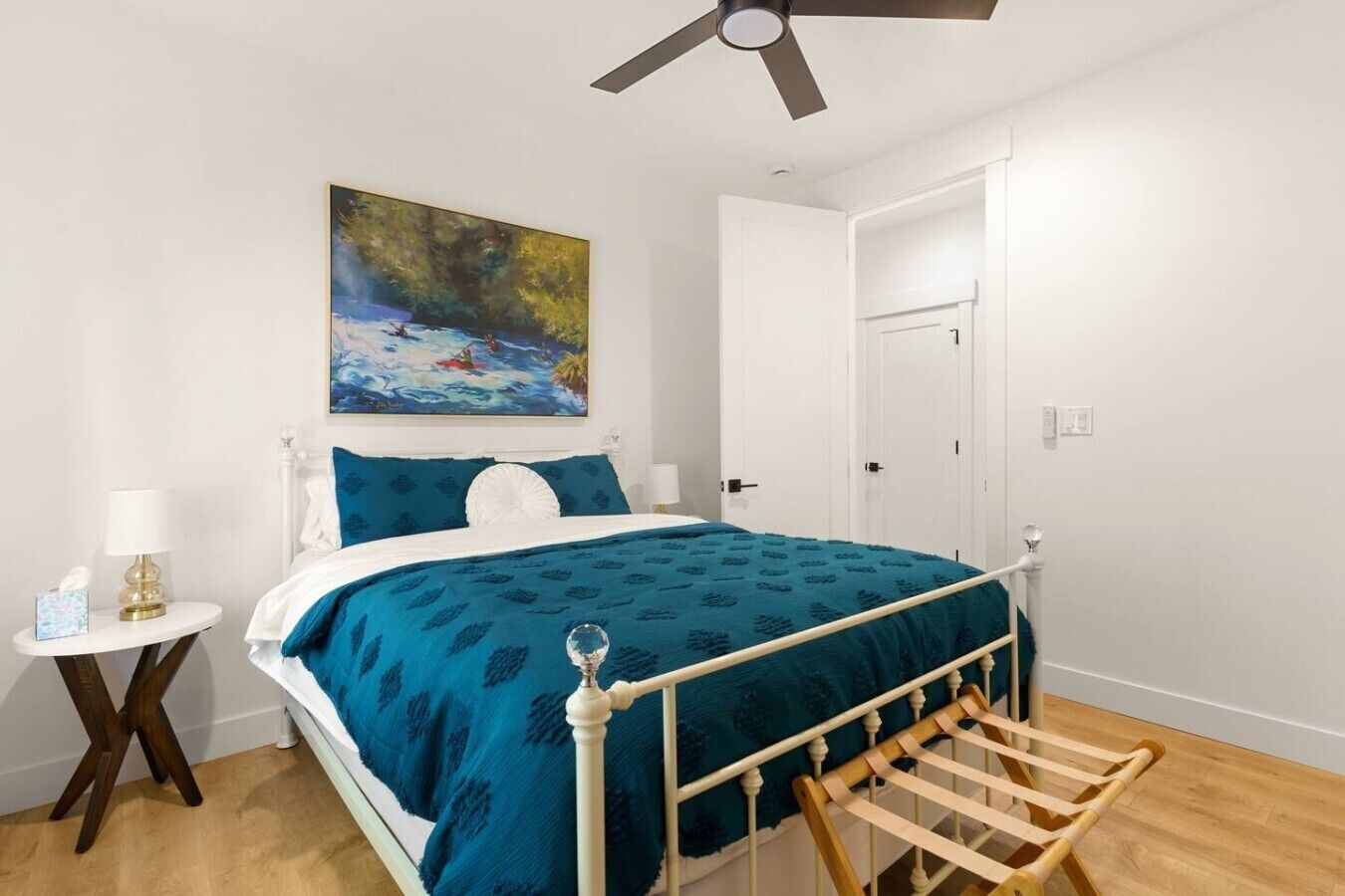 A bedroom with a bed, a painting, a bedside table with a lamp, and a wooden drying rack.