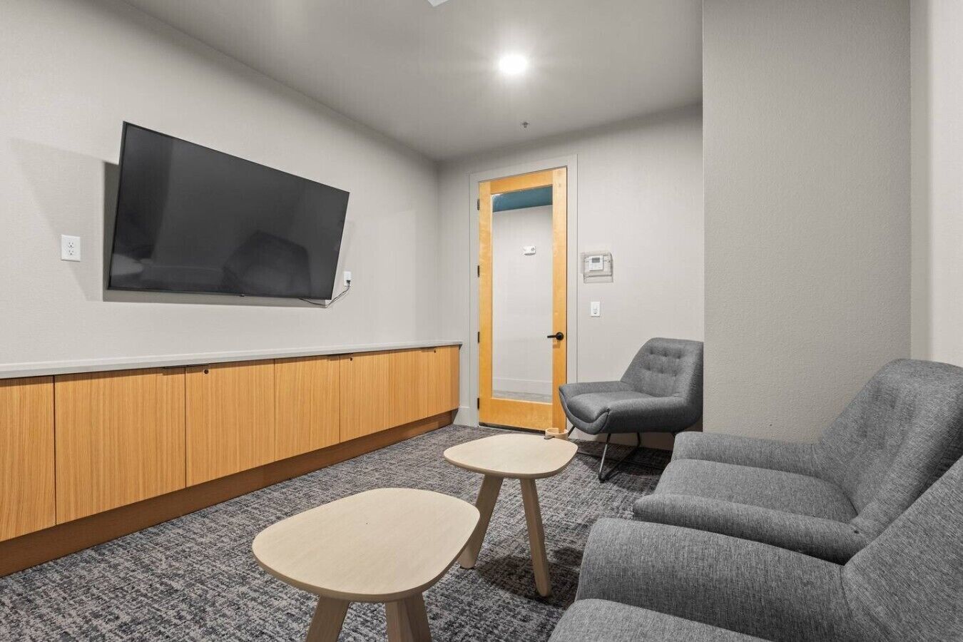 Una habitación con una televisión de pantalla plana montada en la pared, un mueble de madera debajo, un sofá gris, un sillón gris y una pequeña mesa redonda.