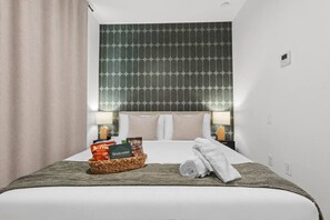 Ein Hotelzimmer mit einem ordentlich bezogenen Bett, einem Willkommensschild, einer Snackkörbchen und einer Nachttischlampe.