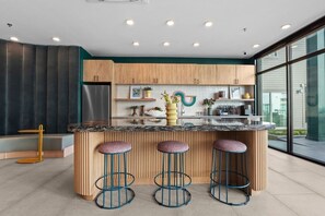 A modern kitchen with a central island, bar stools, wooden cabinets, and a large window providing a view outside. - Luxury Studio Bishop Arts | Coffee Bar | 7 Min DT (Dallas)