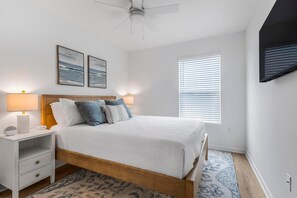 Ein Schlafzimmer mit Bett, Nachttischen, Deckenventilator, Wandbildern, einem Fenster mit Jalousien und einem gemusterten Teppich.