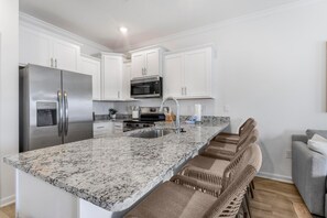 A modern kitchen with white cabinetry, stainless steel appliances, a granite countertop, and a built-in island with bar stools. - Lost Bay Townhome 4239 (Orange Beach)