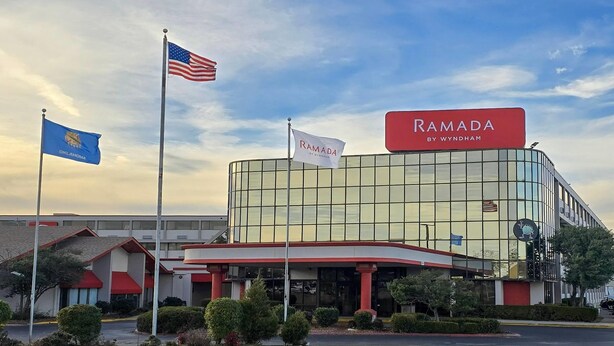 A Ramada hotel with a modern glass facade, a parking lot, and flags of the USA and Oklahoma. - Pet-Friendly Tulsa-Area Stay with Free Breakfast & Parking (Broken Arrow)