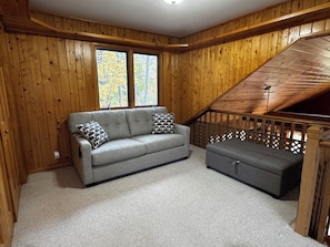 Una habitación con paredes de madera, un sofá gris, un reposapiés a juego y una escalera de madera.