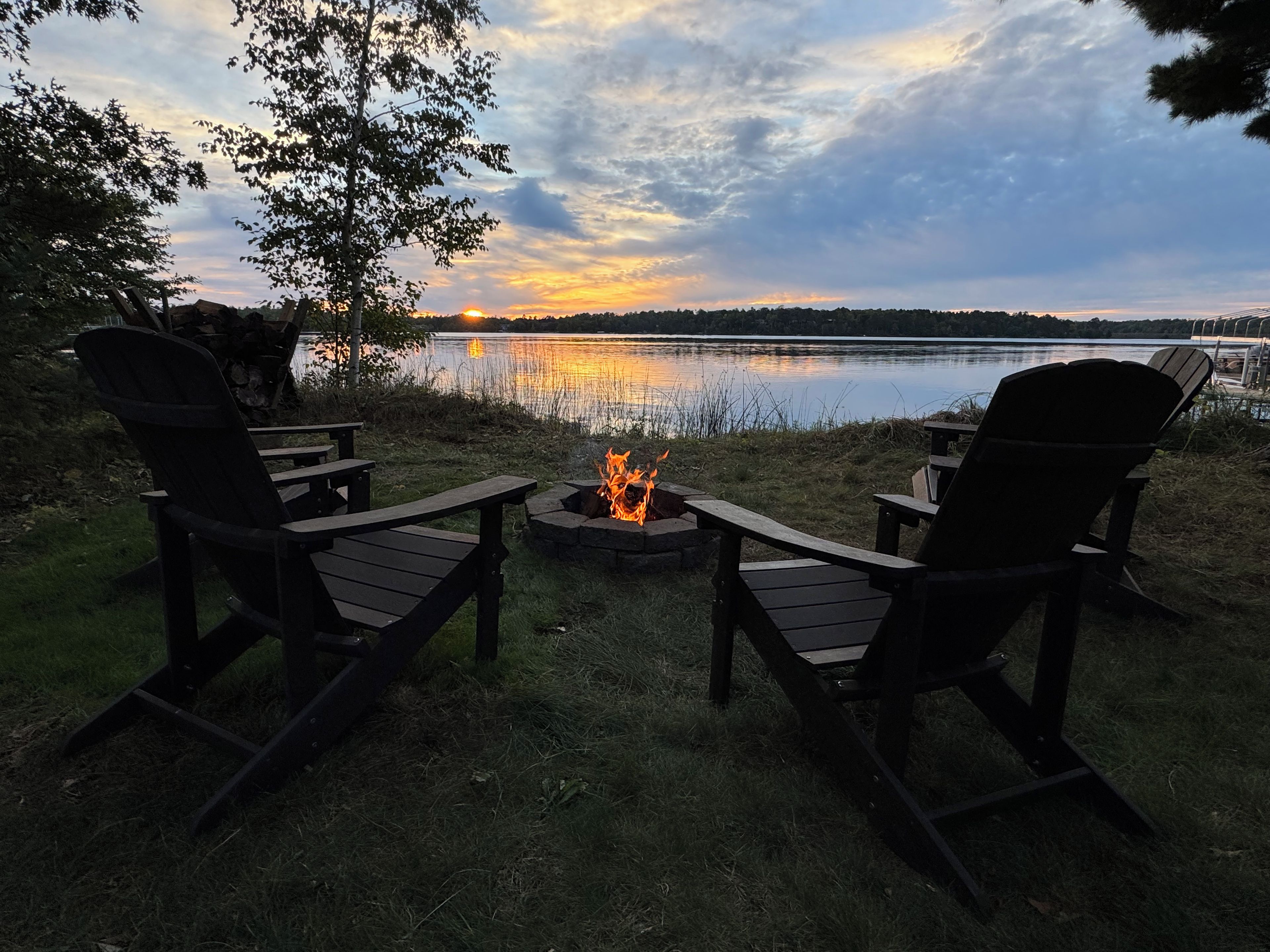 Two Adirondack chairs facing a campfire with a lake and sunset in the background.