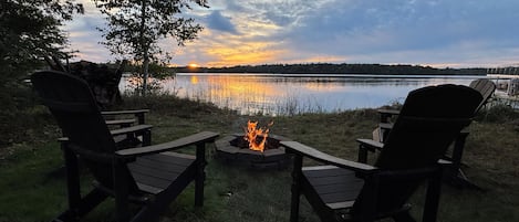 Two Adirondack chairs facing a campfire with a lake and sunset in the background.