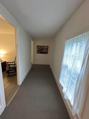 A hallway with a carpeted floor, a painting on the wall, and a window with sheer curtains.