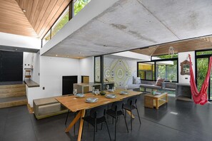 Un espace repas moderne avec une longue table en bois pouvant accueillir six personnes, des chaises noires et un fauteuil suspendu rouge.