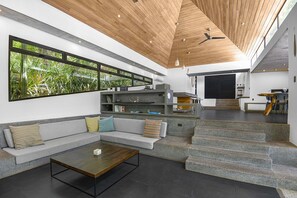 Uma sala de estar ampla com design moderno, destacando uma grande janela, um sofá embutido, uma mesa de centro e uma escadaria que dá acesso a outro nível.