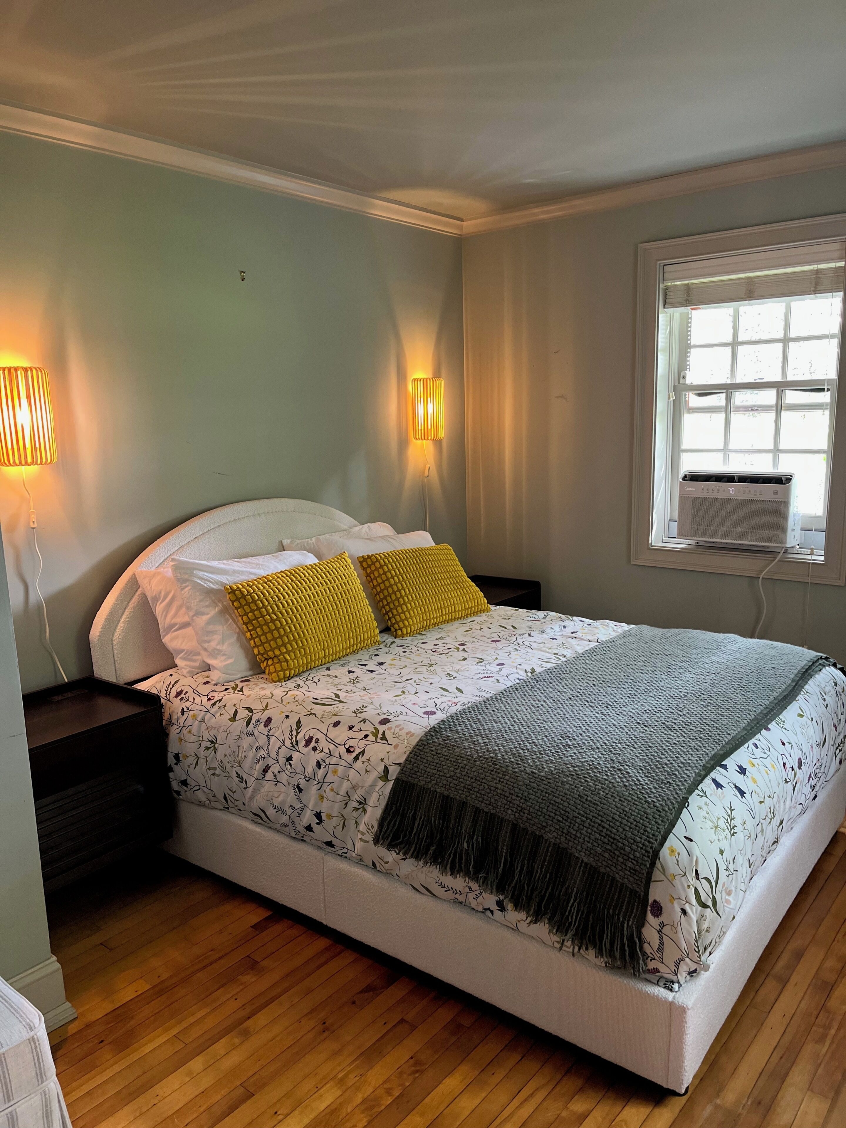 Une chambre à coucher avec un lit, deux lampes de chevet, une fenêtre avec un climatiseur et un plancher en bois.