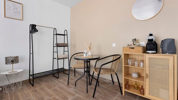 A small, modern room with a round table, two chairs, a ladder, a mirror, a coffee machine, and a cabinet with glassware.