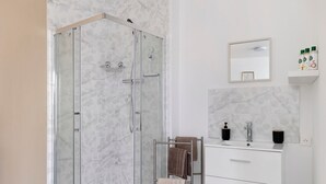 A modern bathroom with a glass-enclosed shower, a white vanity with a mirror, and a small shelf with toiletries.