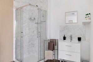 A modern bathroom with a glass-enclosed shower, a white vanity with a mirror, and a small shelf with toiletries. - La Parenthèse du Loir – Guest Room with Private Bathroom and Kitchenette (Montval-sur-Loir)