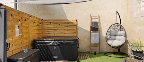 Un espacio exterior reducido con un separador de madera, un jacuzzi, una silla colgante y una mesita con una toalla.