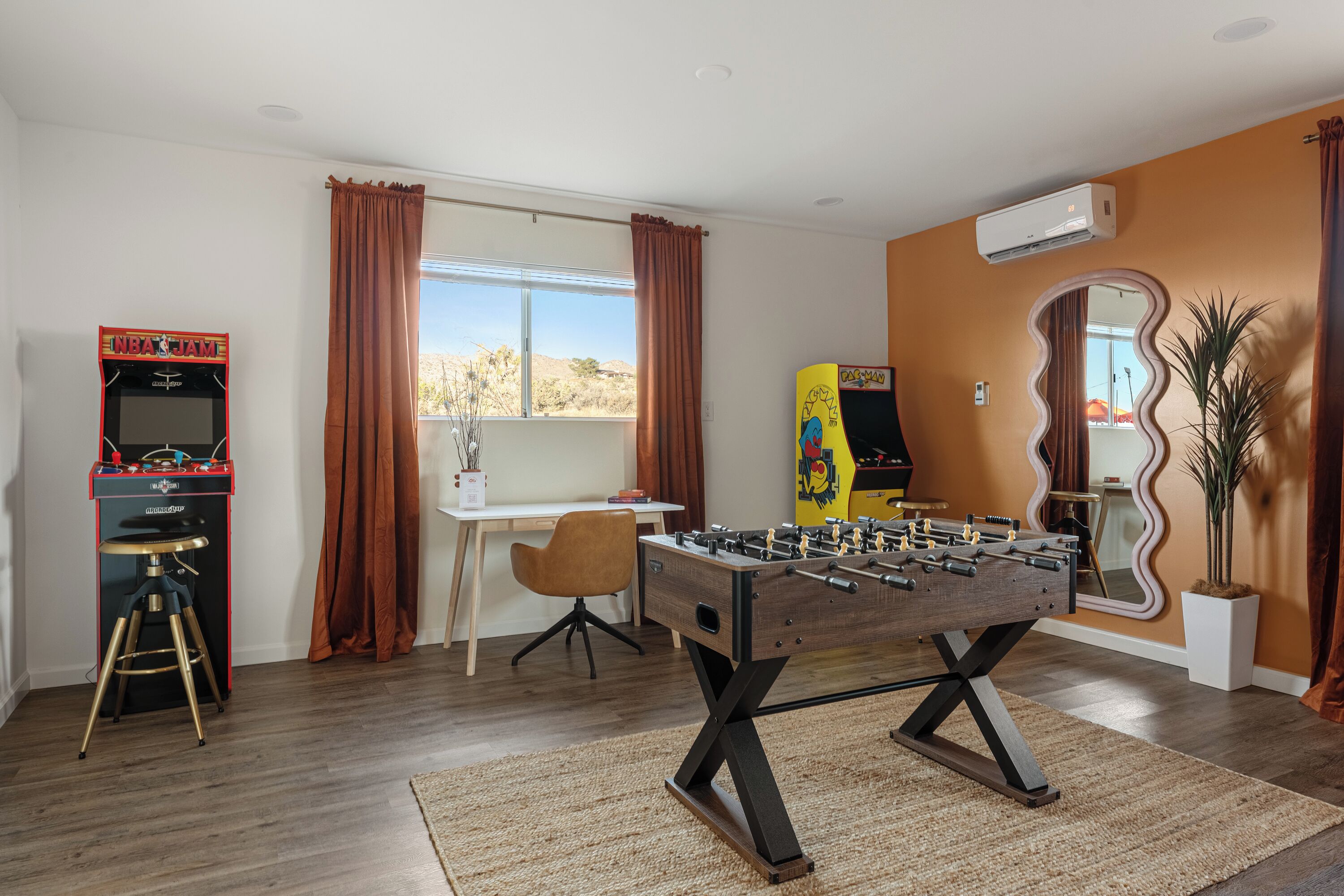 A room with a foosball table, a desk, a chair, a window with curtains, and a mirror on the wall.