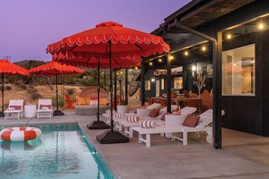 A pool area with lounge chairs and umbrellas, a dining area with tables and chairs, and a building with a dark exterior. - Desert Dreams: private pool, sauna, cold plunge, star dome, gym and theatre (Yucca Valley)