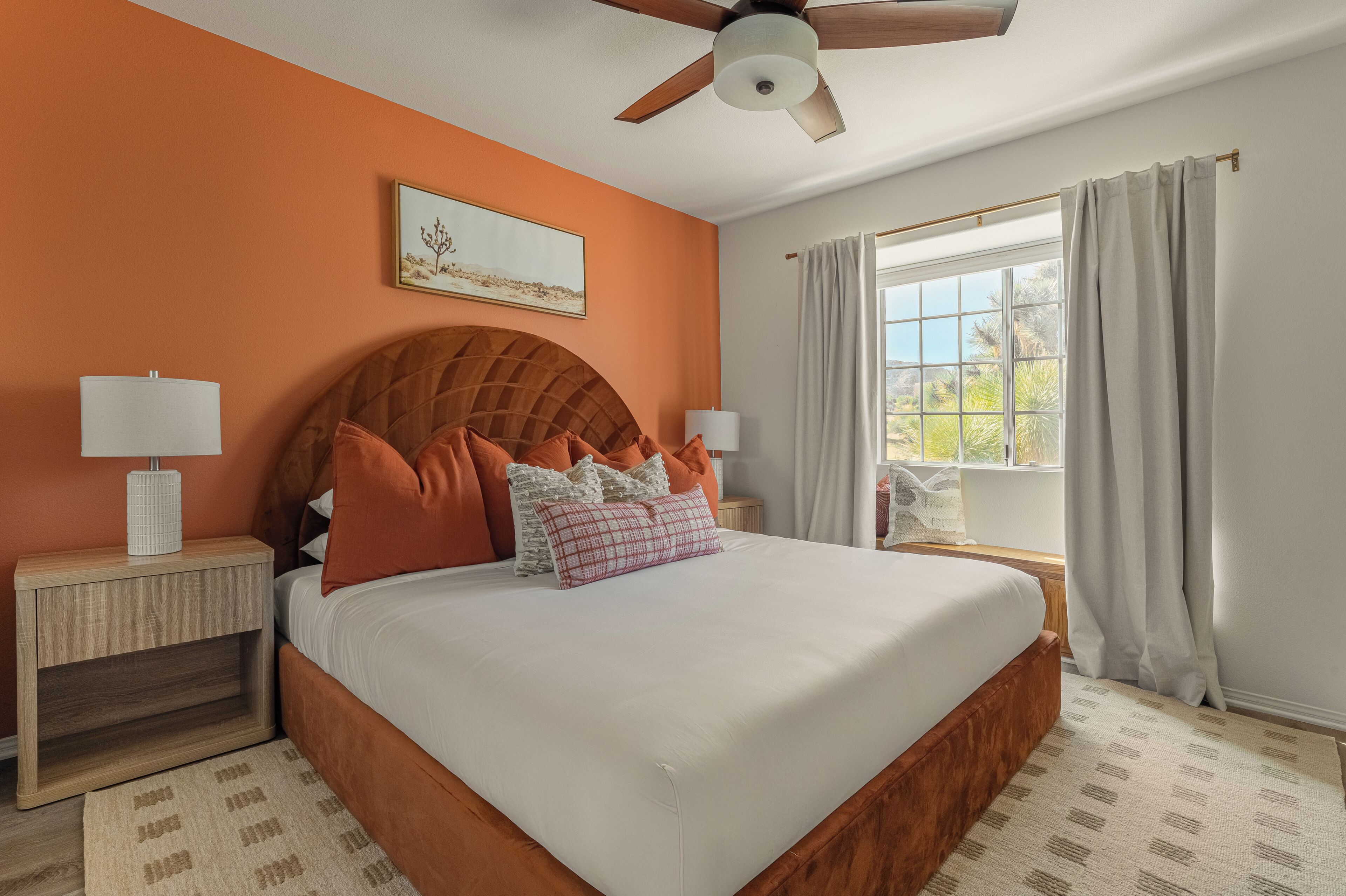 A bedroom with a large bed, wooden headboard, two bedside tables with lamps, a ceiling fan, and a window with curtains.