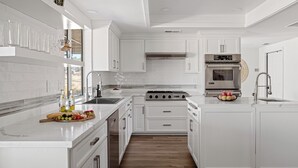 A modern kitchen with white cabinetry, a central island with fruit and bottles, a built-in oven, and a stainless steel range hood. - Desert Dreams: private pool, sauna, cold plunge, star dome, gym and theatre (Yucca Valley)
