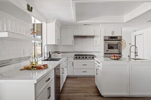 A modern kitchen with white cabinetry, a central island with fruit and bottles, a built-in oven, and a stainless steel range hood. - Desert Dreams: private pool, sauna, cold plunge, star dome, gym and theatre (Yucca Valley)