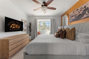 Une chambre avec un grand lit, un ventilateur de plafond, une télévision et une fenêtre avec des rideaux.