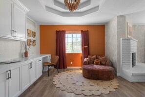 A modern living room with a fireplace, a round ottoman, and a large window with curtains. - Desert Dreams: private pool, sauna, cold plunge, star dome, gym and theatre (Yucca Valley)