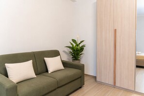 A green sofa with two pillows, a potted plant, and a wooden wardrobe in a room with light-colored walls and flooring. - [Luxury Suite] Ponente Apartment (Giovinazzo(BA))
