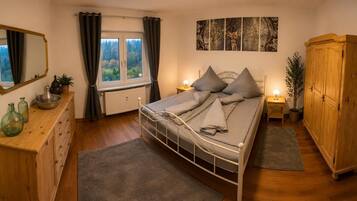 A bedroom with a bed, a wooden dresser, a wardrobe, a window with curtains, and a wall-mounted artwork.