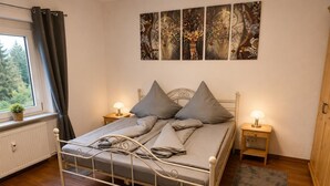 A bedroom with a metal bed frame, grey bedding, two bedside tables with lamps, a radiator, a window with curtains, and a wall with abstract art. - Modern Vacation Rental in Idar-Oberstein (Algenrodt) (Idar-Oberstein)