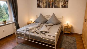 Ein Schlafzimmer mit einem Metallbettgestell, grauer Bettwäsche, zwei Nachttischen mit Lampen, einem Heizkörper, einem Fenster mit Vorhängen und einer Wand mit abstrakter Kunst.