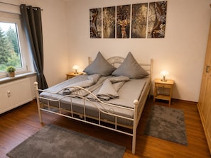 Ein Schlafzimmer mit einem Metallbettgestell, grauer Bettwäsche, zwei Nachttischen mit Lampen, einem Heizkörper, einem Fenster mit Vorhängen und einer Wand mit abstrakter Kunst.