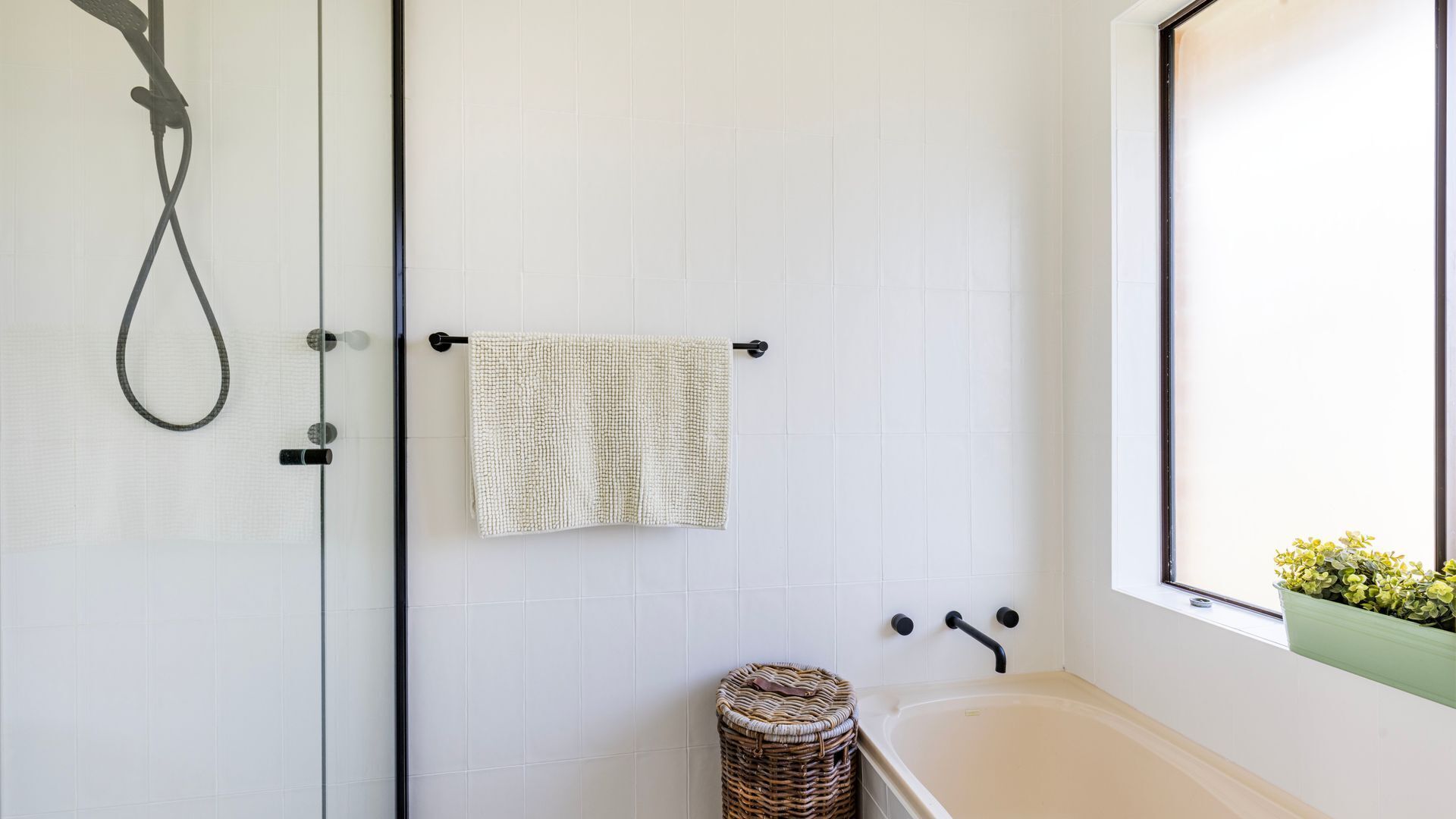 Une salle de bain avec une douche, un porte-serviettes avec une serviette, un petit panier en osier et une fenêtre avec une plante.