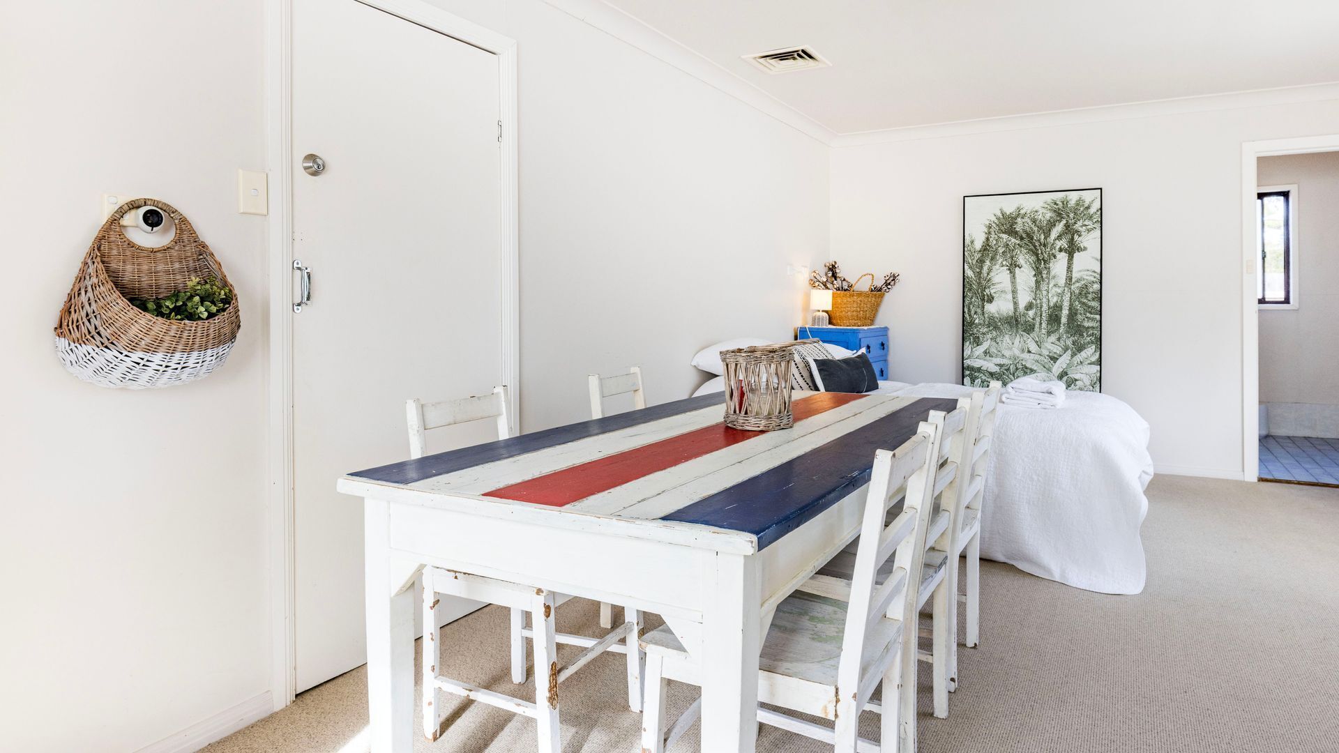 A dining area with a white table and chairs, a woven basket on the wall, and a framed picture on the wall.