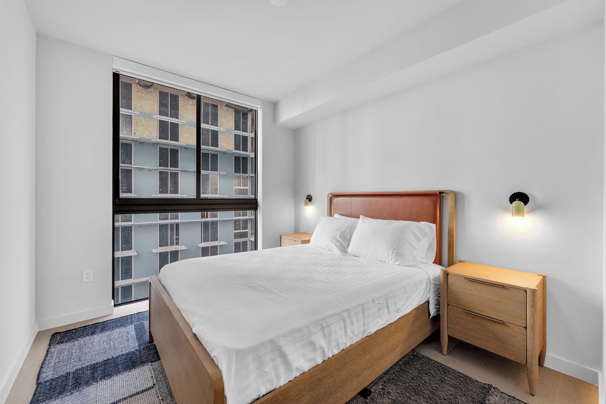 A bedroom with a large bed, a nightstand, a wall-mounted light, and a window with a view of a building.