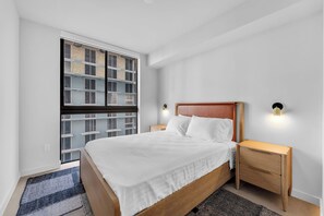 A bedroom with a large bed, a nightstand, a wall-mounted light, and a window with a view of a building. - Chic Downtown Miami 2BR City Views Rooftop Pool (Miami)