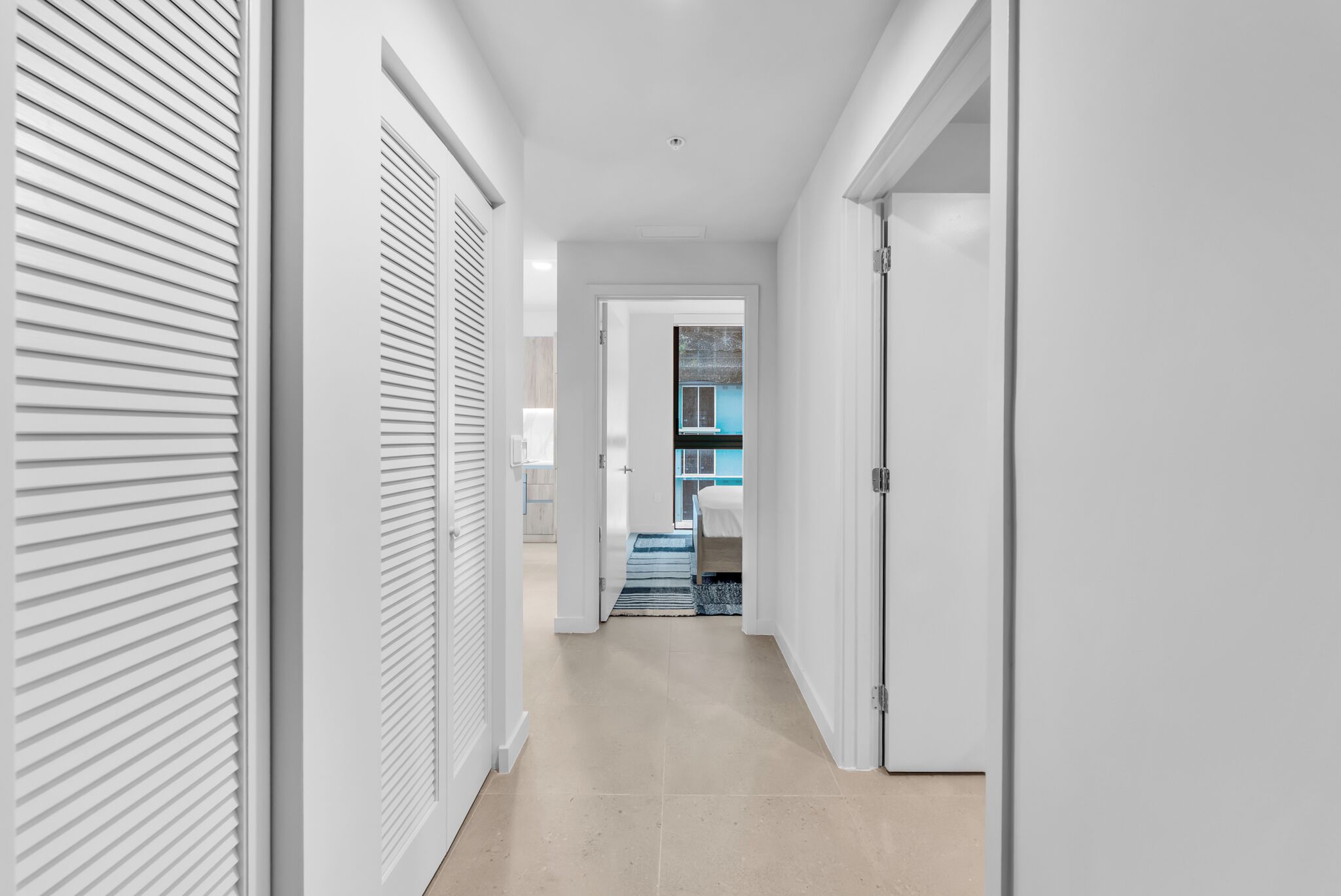 A hallway with white walls, a tiled floor, and a door leading to a room with a blue window.