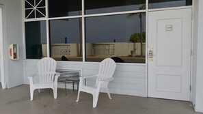 Una puerta blanca con el número 509, una ventana con vista a palmeras y dos sillas blancas con una pequeña mesa frente a ellas.