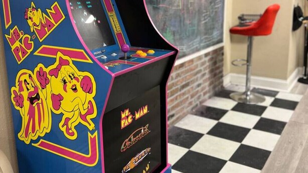 Ein Pac-Man-Automaten in einem Raum mit Schachbrettfußboden und einem roten Sessel.