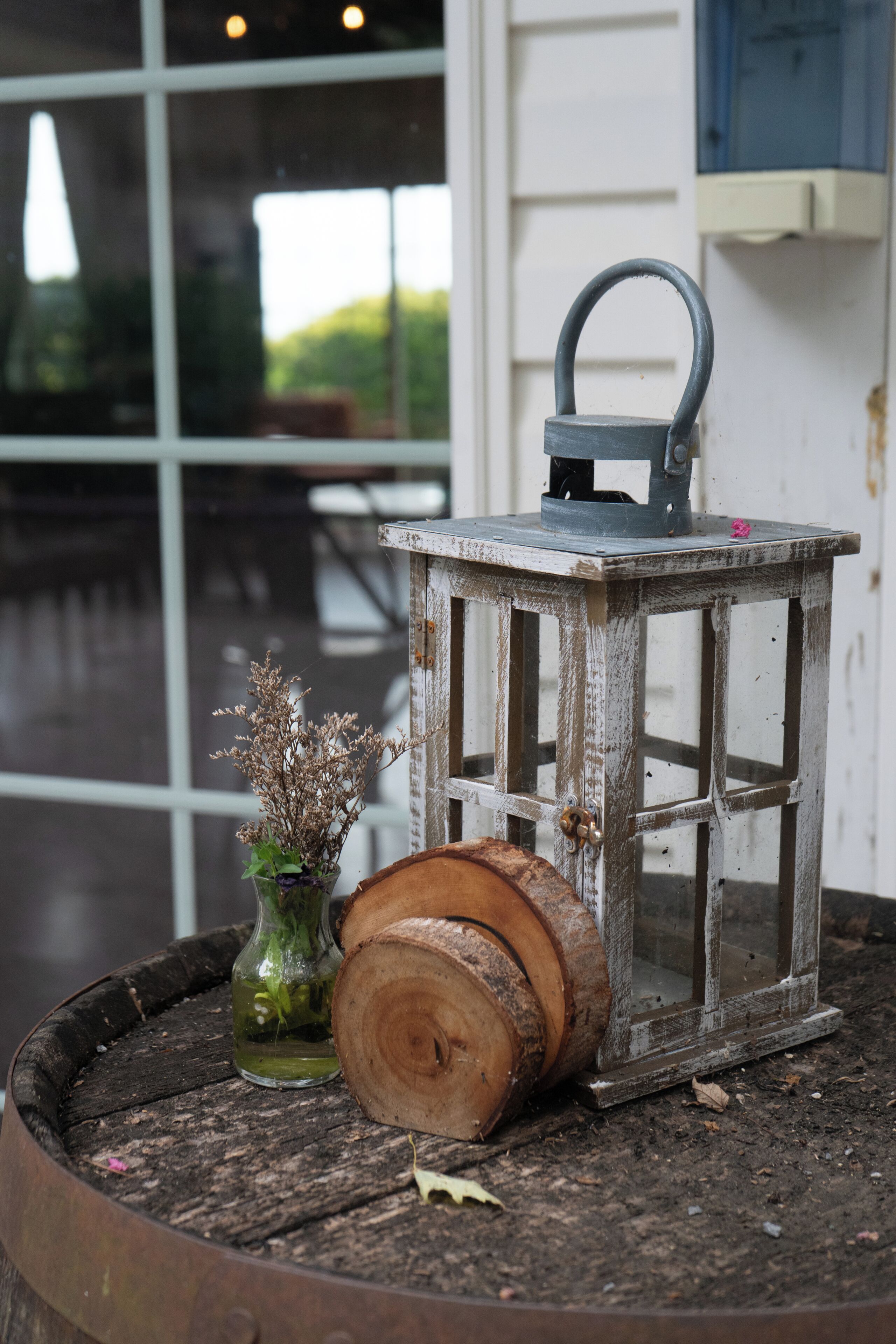 Un farol rústico sobre un barril de madera, un florero de vidrio con flores secas, y una ventana con vista a la vegetación.