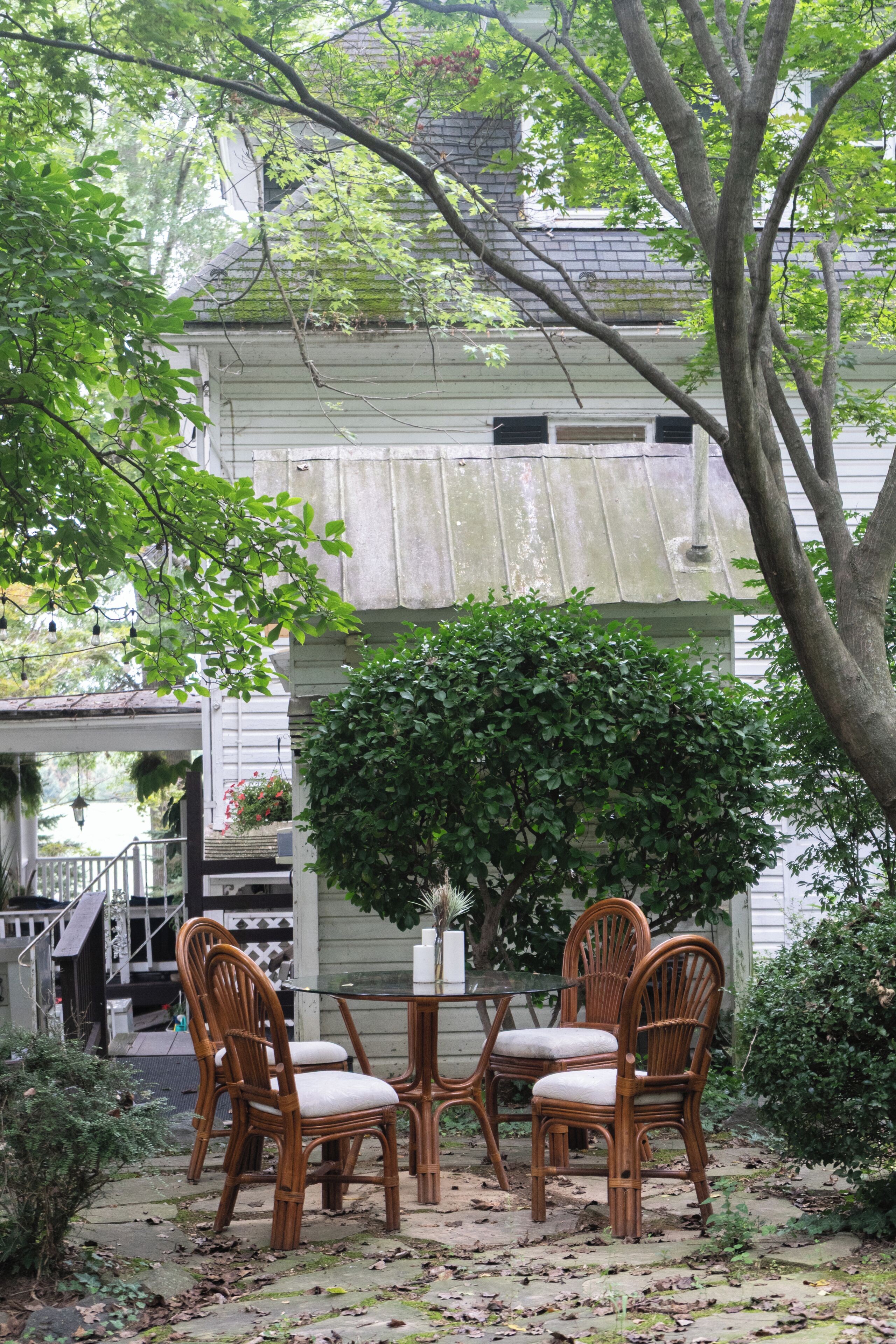 A patio with a table and four chairs, surrounded by greenery and a building with a sloped roof.
