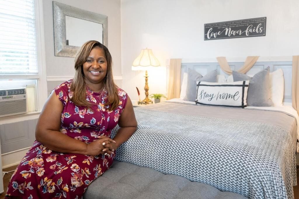A hotel room with a bed, a bedside lamp, a pillow with 'stay home' text, and a wall sign saying 'Love Never fails'.