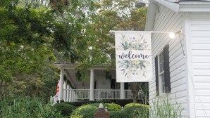 A white house with a porch, a welcome sign, and a garden with a stone marker. - Sunday Morning at Savannahs (New Windsor)