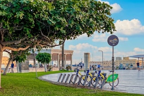 Um parque com uma estação de aluguel de bicicletas, uma árvore grande e um edifício ao fundo.