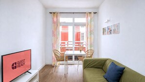 A modern living room with a flat-screen TV, a green sofa, a dining table with chairs, and a view of a building through a window with patterned curtains. - GuestReady - Rosamar M Apartments (Las Palmas de Gran Canaria)