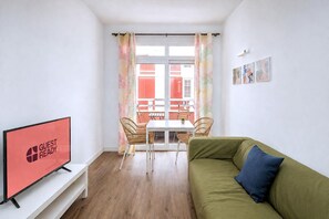 A modern living room with a flat-screen TV, a green sofa, a dining table with chairs, and a view of a building through a window with patterned curtains. - GuestReady - Rosamar M Apartments (Las Palmas de Gran Canaria)