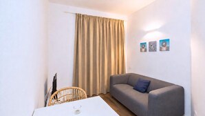 A small, modern living space with a grey sofa, a white table, a wicker chair, and a window with beige curtains. - GuestReady - Rosamar M Apartments (Las Palmas de Gran Canaria)