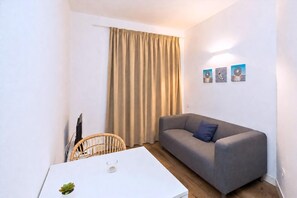 A small, modern living space with a grey sofa, a white table, a wicker chair, and a window with beige curtains. - GuestReady - Rosamar M Apartments (Las Palmas de Gran Canaria)