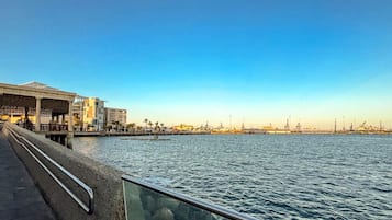 A waterfront promenade with a modern building, a clear sky, and industrial structures in the background.