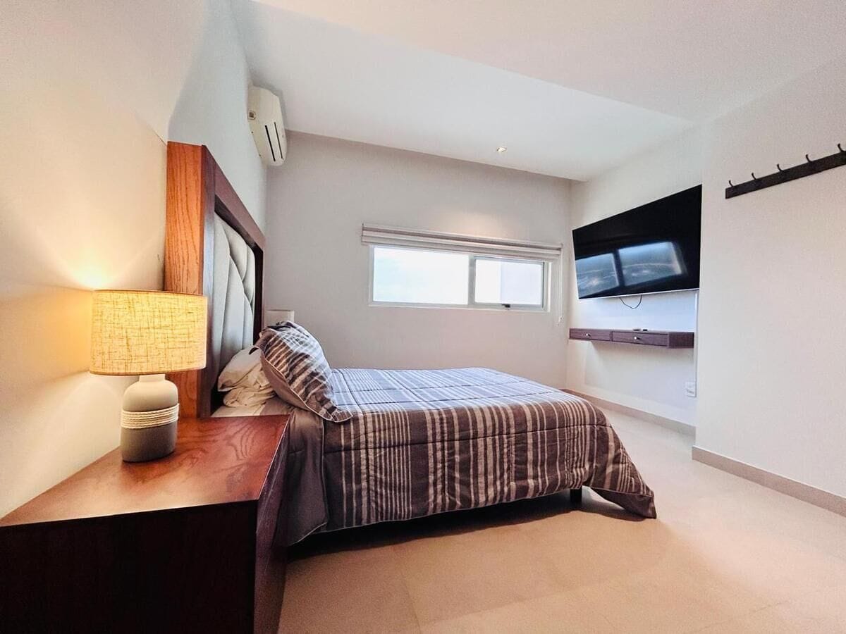 A bedroom with a bed, a bedside lamp, a TV mounted on the wall, and a window.